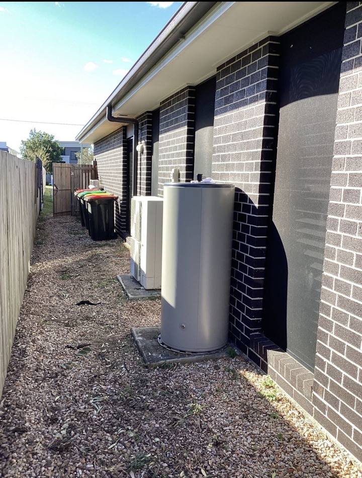 Storage hot water unit standing on a concrete pad in a narrow gravel side yard beside a brick home